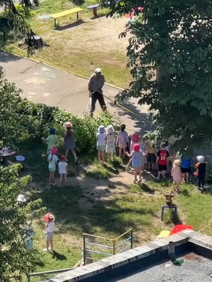 Un jardinero disfruta rociando con agua a un grupo de niños con una manguera, haciéndolos reír, grit…